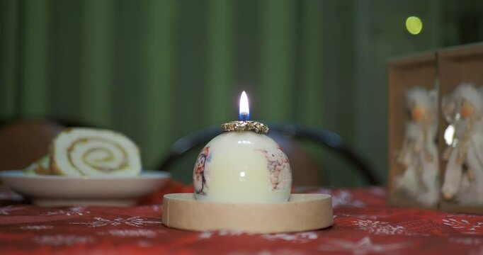 Holiday season. A decorative candle in the form of a ball with painted Christmas pictures stands on a table against a background of flickering multicolored lights. Hand held.