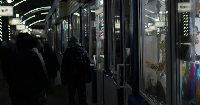 Retail, night city. People in winter clothes walk past the lighted windows of small shops in the evening. Hand held. Siberia.