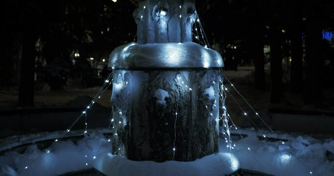 Night city in winter. The stone fountain is decorated with burning iridescent multicolored garlands. Hand held. Siberia.