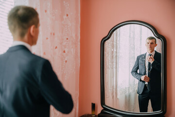 A man in a suit is getting ready for a formal event. He is standing in front of a mirror, adjusting...