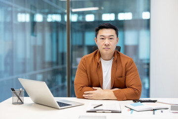 Obraz premium Portrait of confident serious businessman, Asian man looking focused at camera, man behind paperwork with laptop looking at camera.