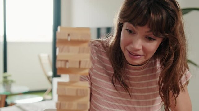 A young woman laughs as she plays Jenga with her partner at home. They're enjoying some quality time playing board games together and the tower almost topples over.