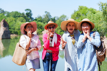 Group of elderly women dressed in casual clothing and wide-brimmed hats, standing near a peaceful lake with a green natural, joyfully chatting, laughing, and enjoying their sunny outing together.
