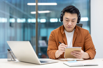 Asian man with headphones and notepad watching educational video course webinar. Man recording data...