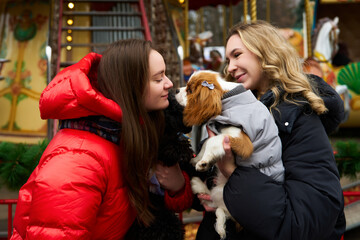 Two friends enjoy a winter day at a fair while playing with their small dogs in cozy jackets