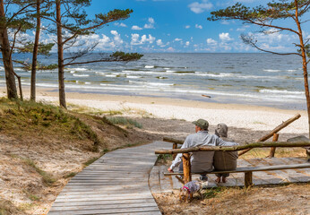 Happy couple of seniors are resting in pine forest and looking at a distance near a sandy beach of the Baltic Sea. Photo depicts ecological tourism in Baltic region. No AI tools were used