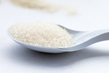 A close-up of granulated white sugar in a white plastic spoon. The sugar crystals are clearly visible. The background is a plain white surface.
