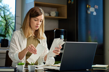 Stressed woman reviewing bills at home. Female organizing receipts, sitting at kitchen table with...