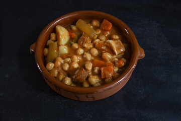 Chickpea stew, served in a clay dish. Dark background. Spanish food. Bacon, chorizo, carrot, meat, vegetables.
