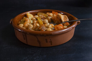 Chickpea stew, served in a clay dish. Dark background. Spanish food. Bacon, chorizo, carrot, meat, vegetables.