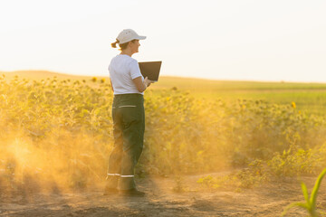 Woman farmer wearing white cap and t-shirt with laptop on agricultural field at sunset.