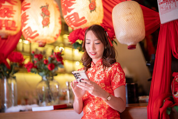 A woman in a red Chinese dress is sitting at a bar and looking at her cell phone. The scene is set in a restaurant with red lanterns hanging from the ceiling