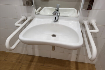 Wall-mounted white sink with chrome faucet and safety grab bars on both sides. Bathroom interior with mirror and tiled walls. Accessibility design for public restrooms.