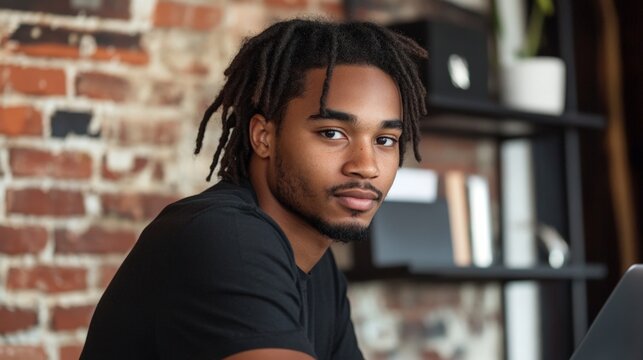 A man with dreadlocks is sitting in front of a bookshelf. He is wearing a black shirt and has a beard
