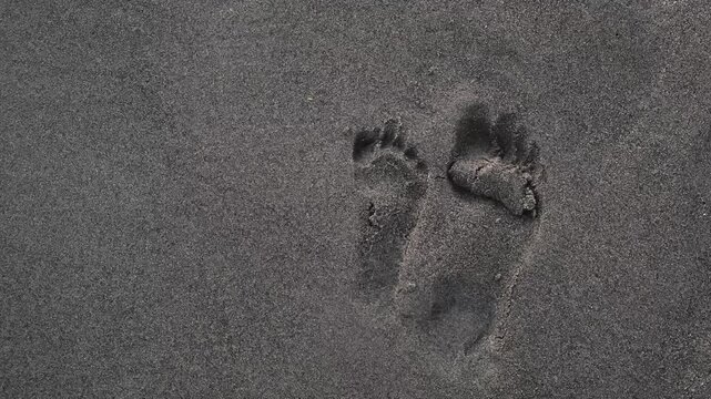 Sea waves washing off pair of big and small human footprint from volcanic sand. Traces of a child's and an adult's foot. End of family beach holiday. The concept of the frailty of human existence. 4K