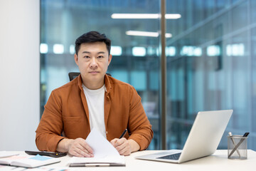 Fototapeta premium Portrait of serious successful financier investor. Man behind paperwork looking at camera with concentration. Businessman working with laptop and documents inside office.
