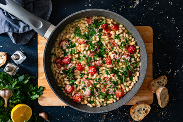 White beans with cherry tomatoes, parmesan, lemon and garlic in frying pan on black table