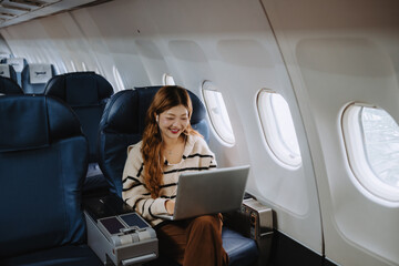 Asian businesswoman wearing earphones, working on a laptop while seated by the window in an airplane, enjoying a comfortable and productive business trip during her journey