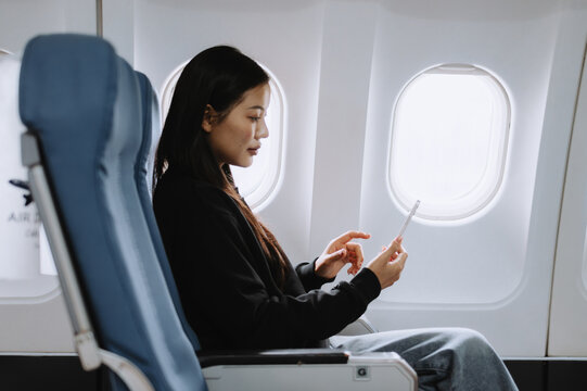 Young asian businesswoman using her smartphone during a flight, enjoying the convenience of technology while traveling, seated comfortably in an airplane near the window