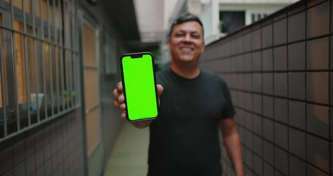 Man smiling while holding smartphone with green screen and chroma key towards camera, showcasing customization or mockup potential in a home corridor