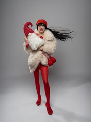 A young, beautiful woman in a white fashionable fur coat, red tights, and a red beret, holding a Finnish Santa toy. She has stunning black hair, adding elegance to the festive look.
