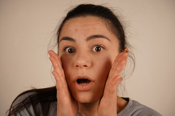 The expression vividly captures a moment of astonishment and surprise. Her pose emphasizes the unexpected reaction. A young woman places her hands to her cheeks, her eyes wide open in genuine shock.