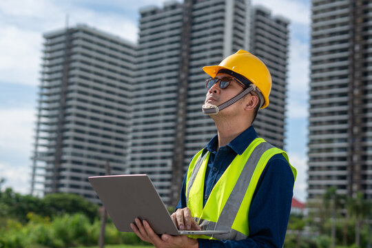 A construction worker uses a laptop while surveying a modern urban skyline.