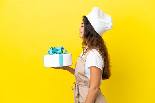 Young caucasian pastry chef woman with a big cake isolated on yellow background laughing in lateral position
