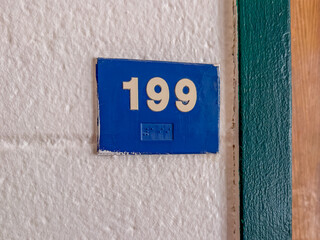 Blue 199 sign with white lettering and braille, on a blue block wall.	