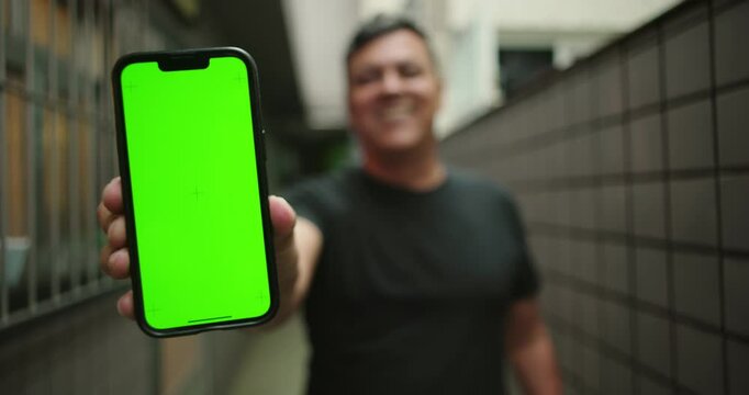 Man reacts positively to content on smartphone, smiling warmly before presenting phone with green screen and chroma key to camera in an outdoor home corridor