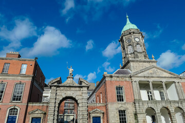 Dublin Castle's Bedford Tower in Ireland. Green dome and clock faces, stands prominently against blue sky. Stone gateway topped by statue of Justice leads into castle grounds. Georgian brick buildings