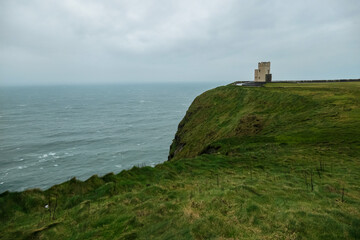 Dramatic coastal scene of Cliffs of Moher in Ireland. Steep cliff drops directly into Atlantic Ocean. Choppy sea with white waves crashing against base of rock formation. Iconic O'Brien's Tower on top