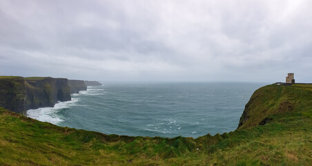 Dramatic coastal scene of Cliffs of Moher in Ireland. Steep cliff drops directly into Atlantic Ocean. Choppy sea with white waves crashing against base of rock formation. Iconic O'Brien's Tower on top