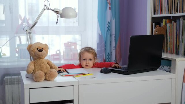 Cute baby girl sitting at workdesk with laptop in nursery, teddy bears and toys nearby. Freelance and distance work concept