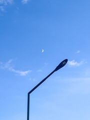 Moon peeks above street lamp during clear day