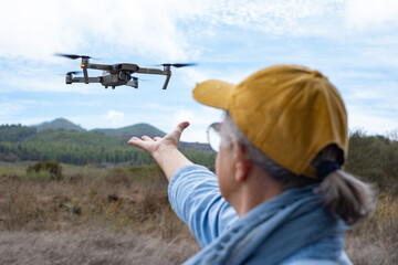 Rear view of an elderly woman flying a drone in a forest area using the remote control. The drone pilot reaches out to pick up the drone. Aerial nature photography and videography