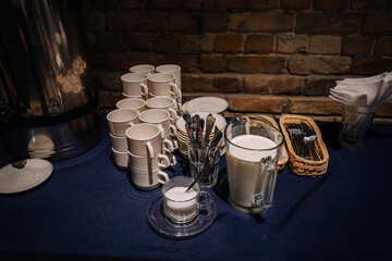 Table with stacked white cups, plates, milk jug, sugar bowl, and utensils in baskets, set against a brick wall background, ready for serving..