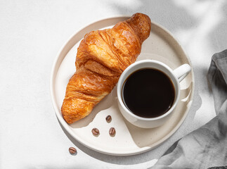 Cup of espresso coffee and fresh croissant on white plate on light textured background with shadow and napkin