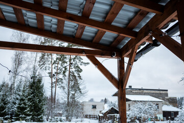 A wooden terrace with a corrugated metal roof and string lights, set against snow covered trees,...
