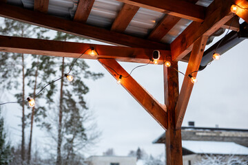 Obraz premium A wooden terrace with string lights glowing warmly, corrugated metal roof, tall trees, snow covered ground, and a modern flat roof building in view.
