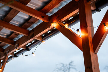 Outdoor wooden terrace structure with a corrugated metal roof, featuring warm glowing string lights...