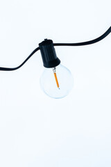 A single LED bulb with visible orange filament hangs from a black wire, set against a bright, overcast sky in a minimalistic composition.