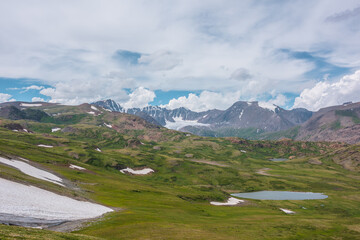Fototapeta premium Scenic awesome landscape with big snowfield and alpine lake among green hills and rocks with view to glacier and large snow-capped mountain top far away under clouds in blue sky in changeable weather.