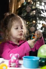 Fototapeta premium The child plays with toys. A little girl sits at a table, against the backdrop of a Christmas tree. Toys are located on the table.