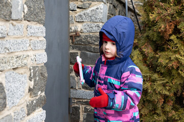 A little girl stands near a stone wall and curiously explores the surroundings. The child is dressed in a warm winter jumpsuit. The child holds a toy in her hand.