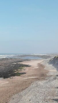 DRONE FOOTAGE - Sea, beach boulders, pebble shore and waves. Sea waves breaking on rocks. Sunny beach with sand dunes and blue sky in Esposende, Portugal. Spring Wind Day.