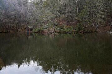 Small shed on the shore of a calm lake with trees reflected. 