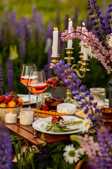 Elegant gorgeous wedding table decor or romantic dinner arrangement outdoors in blooming field. Purple lupine flowers, candles, fruits and wine, wooden vintage furniture. Sunset, summer, golden hour