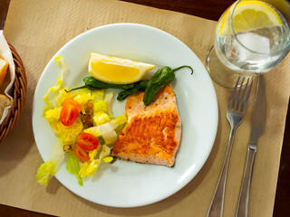 Delicious baked salmon fillet served with fresh vegetable salad, slice of lemon and roasted jalapenos..
