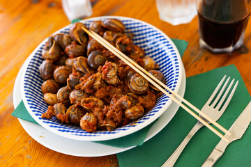 On wooden table there is plate filled to brim with boiled snails. Shellfish snail cooked with minced meat and tomato dressing. Chopsticks are served with food.
